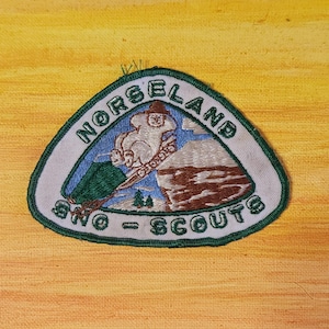 May include: A triangular embroidered patch with a dark green border featuring the text NORSELAND SNO-SCOUTS. The design depicts a white figure skiing down a snowy slope with brown rock formations, a green pine tree, and a blue sky background.