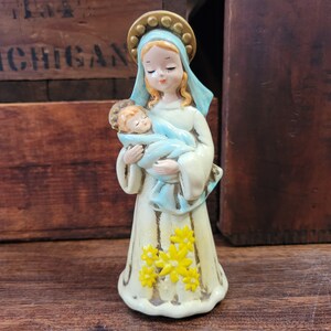 May include: A ceramic figurine of Mary holding baby Jesus, both with closed eyes. Mary wears a white robe with yellow flowers and a blue veil. The figurine has a gold halo and is set against a wooden background.