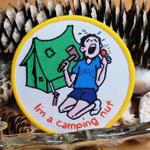 May include: Embroidered patch with a yellow border and a white background. The patch features a cartoon of a person kneeling with a mallet and a lit match, with a green tent in the background. Text reads "I'm a camping nut."