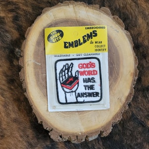 May include: Embroidered patch with the text "GOD'S WORD HAS THE ANSWER" and an image of a hand holding a book with a cross. The patch is in a yellow and clear plastic package. The package reads "EMBLEMS to WEAR COLLECT IDENTIFY".