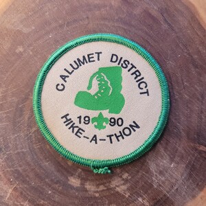 May include: Round patch with green trim and text. The patch reads "CALUMET DISTRICT HIKE-A-THON 1990" with a green hiking boot and a green trefoil symbol. The patch is tan with black and green text.
