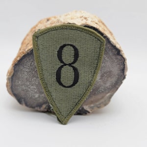 May include: Olive green embroidered patch with the number 8 in black. The patch has a shield shape with a stitched border. The patch is resting on a light brown and gray stone.