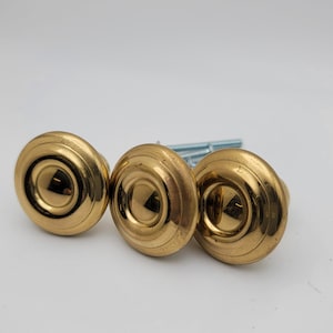 May include: Three gold-colored cabinet knobs with a circular design and a central indentation. The knobs are made of metal and have a shiny finish. Several silver screws are visible behind the knobs. These knobs are suitable for furniture or cabinet hardware.