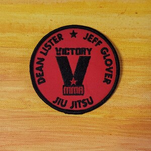 May include: A circular embroidered patch with a red background and black border. It features the black text DEAN LISTER, JEFF GLOVER, and JIU JITSU around the edge. A large black letter V is in the center with the word VICTORY above and MMA below.