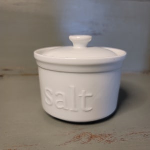 May include: A white ceramic salt cellar with a lid and a small handle on top. The word "salt" is embossed on the front of the container. The salt container is round and sits on a gray surface.