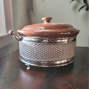 May include: A vintage casserole dish with a brown ceramic lid and a silver-colored metal base. The base has a woven pattern and small feet. The dish has a handle on each side. A classic piece for serving or cooking.