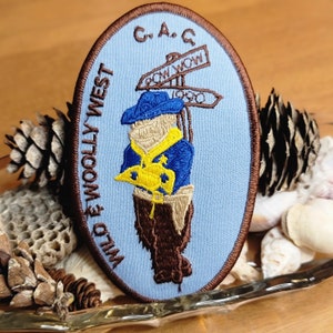 May include: Embroidered patch with a light blue oval background featuring a cowboy figure in blue and brown attire, holding a yellow object. The patch includes the text "WILD & WOOLLY WEST" and "C.A.C." with directional signs that read "POW WOW" and "1990".