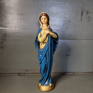 May include: A vintage statue of the Virgin Mary. The figure is depicted with a serene expression, wearing a blue robe and a cream-colored veil. Her hands are clasped in prayer, and the base is a light tan color.