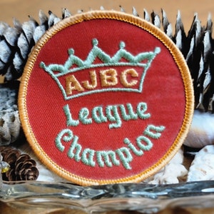 May include: A round, red patch with gold trim and embroidered text. The patch features a crown design above the letters "AJBC" and the words "League Champion" in a script font. The patch is likely a sports or achievement emblem.