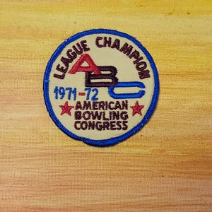 May include: A circular embroidered patch with a cream background and a blue border. It features the text LEAGUE CHAMPION, 1971-72, and AMERICAN BOWLING CONGRESS in dark red and blue. The letters ABC are prominently displayed in red, black, and blue.