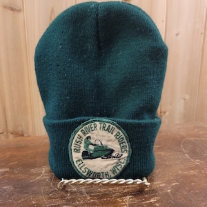 May include: A dark green knit beanie with a folded brim. The beanie features a circular patch with the text "Rush River Trail Riders Ellsworth, Wisc" and an image of a snowmobile. The hat is displayed on a gold-colored stand.