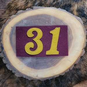 May include: A burgundy felt patch with the number 31 in gold. The numbers are stitched onto the patch. The patch is displayed on a wooden slice with a brown and tan fur-like background.