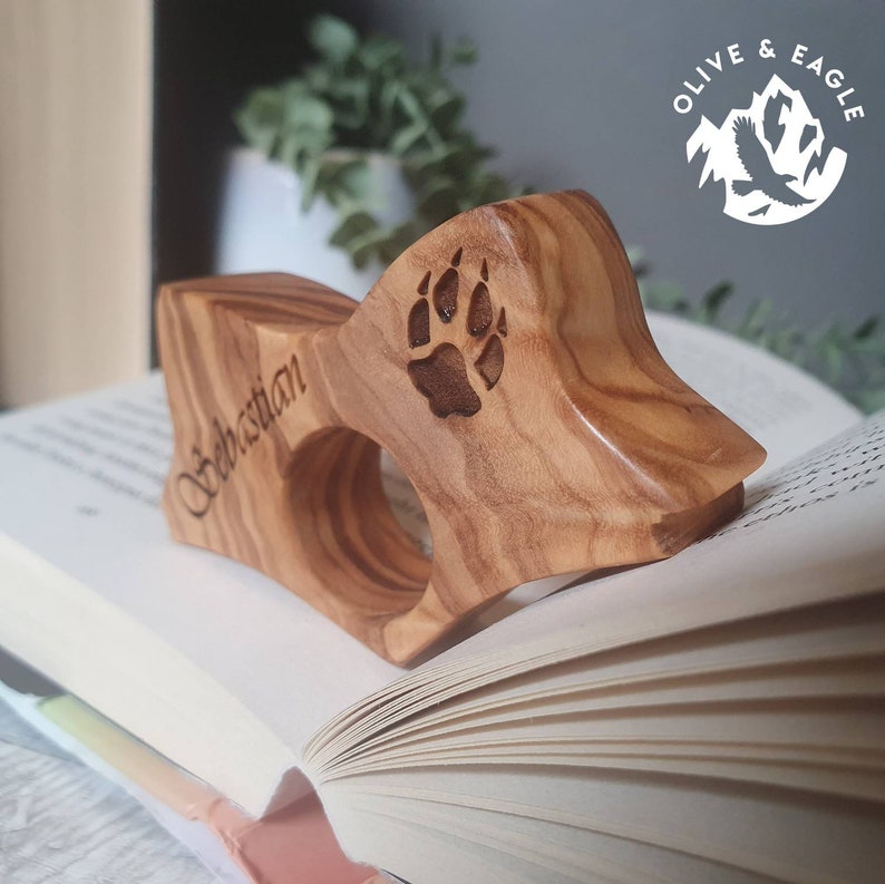 May include: A wooden book holder with a paw print and the name "Sebastian" engraved on it. The holder is made of light brown wood and is resting on the pages of an open book. The logo "Olive & Eagle" is in the background.