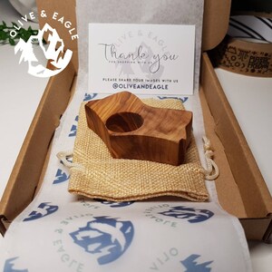 May include: A handcrafted wooden candle holder with a natural wood grain finish, presented in a cardboard box with a thank you card. The candle holder is nestled in a small burlap bag, and the box is lined with patterned tissue paper.