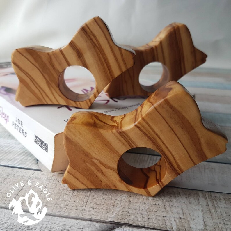 May include: Three wooden book page holders with a star-like shape and a circular opening. The holders are made of olive wood, displaying a natural wood grain pattern. The holders are resting on a book.