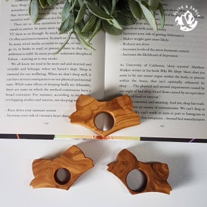 May include: Three wooden fox-shaped book page holders. Each holder has a circular opening and is made of light brown wood with visible grain. The holders are arranged on an open book with text and a sprig of greenery.