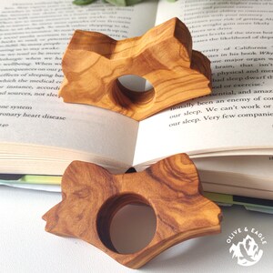 May include: Two wooden fox-shaped book holders with a circular opening, resting on the pages of an open book. The holders are made of light brown wood with visible grain patterns. The book is open to a page of text.
