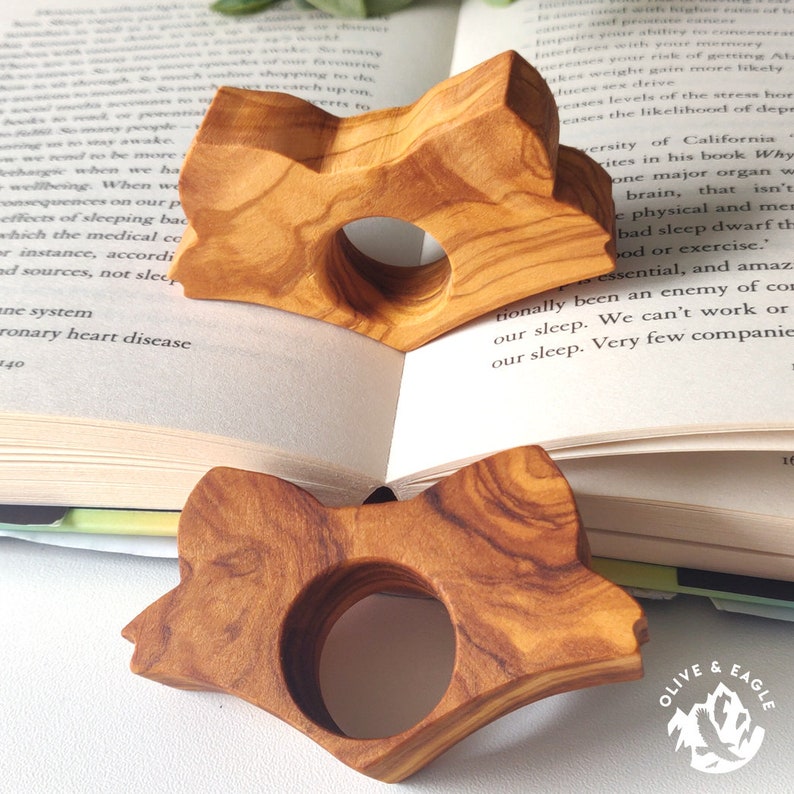 May include: Two wooden fox-shaped book holders with a circular opening, resting on the pages of an open book. The holders are made of light brown wood with visible grain patterns. The book is open to a page of text.