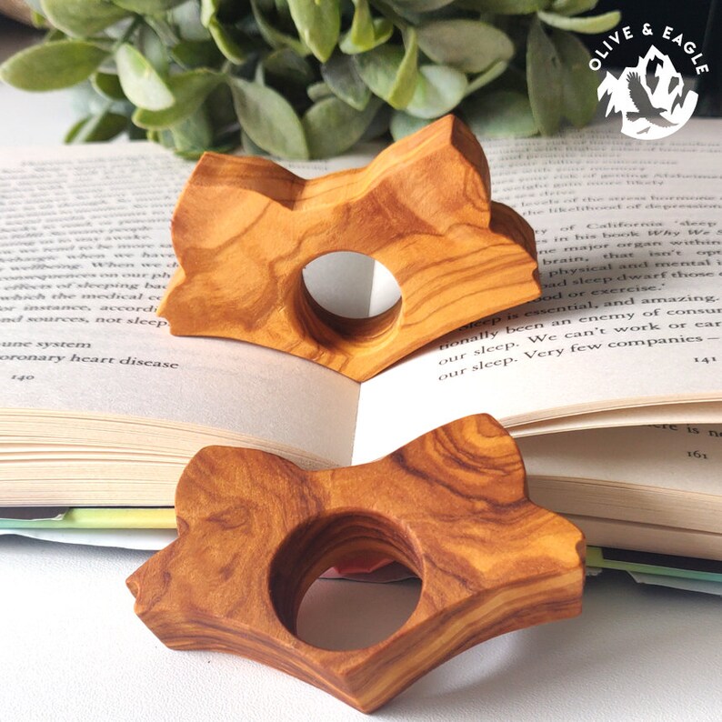 May include: Two wooden fox-shaped book page holders with a natural wood grain pattern. The holders are placed on an open book with text visible. The logo "OLIVE & EAGLE" is in the upper right corner.