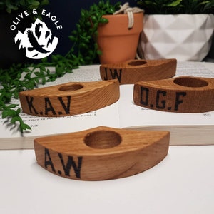 May include: Four wooden ring holders with the initials K.A.V, J.W, D.G.F, and A.W engraved on them. The holders are made of light brown wood and are sitting on a book.