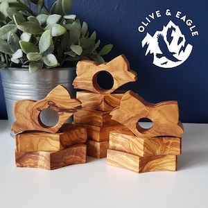 May include: A set of olive wood napkin rings and matching square bases. The napkin rings are shaped like stylized leaves with a circular opening. The wood grain is visible, showing brown and tan tones. The logo "OLIVE & EAGLE" is visible in the background.