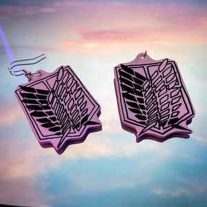 May include: A pair of pink earrings featuring a black emblem design. The earrings have a shield shape with a black wing and rectangular design. The earrings are hanging from silver-colored fish hook ear wires. The background is a reflection of a colorful sky.