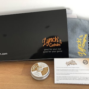 以下が含まれることがあります： 「Lynch's Cue Balm」と黒と金色のラベルが貼られた小さなキューバームの缶。バームは、同じロゴとウェブサイトアドレス「cuebalm.com」が書かれた黒い箱に入っています。同じロゴの灰色の布も含まれています。