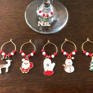 May include: Set of six gold-tone Christmas-themed wine glass charms. Each charm features a different design: reindeer, Santa Claus, mitten, snowman, Christmas tree, and the word "NOEL".