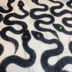 May include: A collection of black beaded snake appliqués with shimmering details and jewel-toned eyes. The snakes are arranged in a pattern, displaying their curved forms and intricate beadwork. These decorative appliqués are suitable for embellishing clothing or accessories.