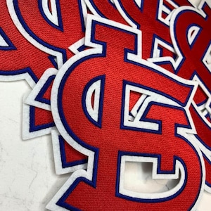 May include: Red embroidered patches in the shape of the letter "S" with a white border and blue outline. The patches are stacked, showing the texture of the fabric and the detailed stitching.