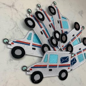 Op de afbeelding: Geborduurde patches in de vorm van een postwagen. De patches zijn wit met blauwe ramen, rood-blauwe strepen en zwarte wielen. De patches hebben een kleine groene lus aan de bovenkant.