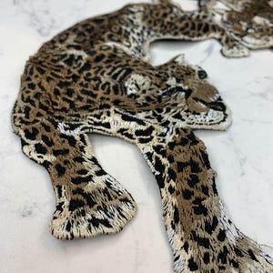 May include: Embroidered leopard applique in shades of brown, black, and cream. The applique features a detailed design of a leopard with its body and legs extended. The leopard's spots are visible, and the edges are neatly stitched.