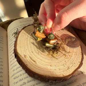 May include: Handmade frog earrings. Each earring features a small, green frog perched atop a stack of miniature books. The books are in shades of brown and orange. The earrings are held up by gold-colored hooks.