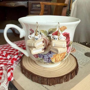 May include: A pair of dangle earrings featuring miniature stacks of two beige books topped with a small white teapot and a white teacup with black details. The earrings are suspended from gold-toned metal hooks and are designed as charms.