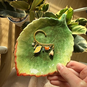 May include: A gold-toned ear cuff with a leaf, butterfly, and flower charm. The ear cuff is displayed on a green leaf-shaped ceramic dish with a red and yellow rim. A potted plant is in the background.