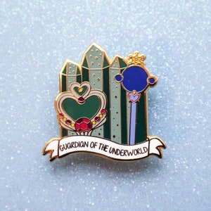 May include: Gold enamel pin featuring a green heart with a red gem, a blue orb on a stick, and a banner that reads "Guardian of the Underworld".