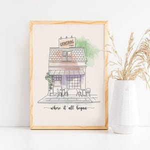 May include: A framed print featuring a line drawing of a cafe with the text "CENTRAL & CAFE" on a sign. The image includes outdoor seating and the phrase "where it all began". The artwork is in a light wood frame.