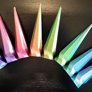 Puede incluir: Una colección de siete objetos puntiagudos geométricos multicolores dispuestos en semicírculo. Los colores incluyen morado, rosa, rojo, naranja, verde, azul y azul claro. Los objetos tienen un brillo metálico.