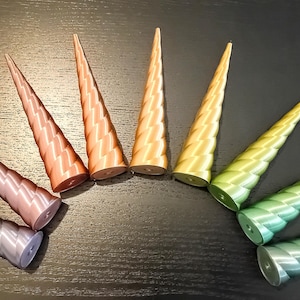 Puede incluir: Una variedad de objetos cónicos coloridos con un patrón en zigzag. Los objetos son de color plata, rosa, cobre, oro, verde y amarillo. Los objetos están dispuestos en semicírculo sobre una superficie oscura.