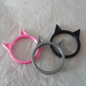 May include: Three cat ear rings in pink, gray, and black. The rings are made of plastic and have a cat ear design.