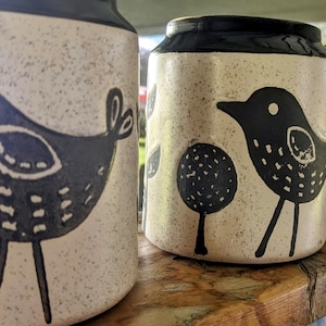 May include: Three ceramic jars with a speckled, off-white finish and black bird designs. The birds have dotted details and stand on thin legs. The jars have black rims and are displayed on a wooden shelf.