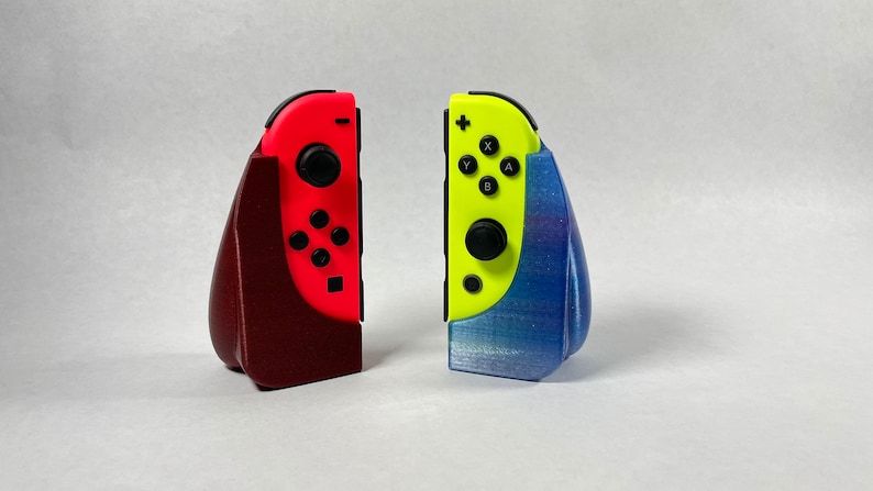 May include: Two Nintendo Switch Joy-Con controllers in red and yellow, each in a 3D printed stand. The red stand is maroon and the yellow stand is blue with a glittery finish.