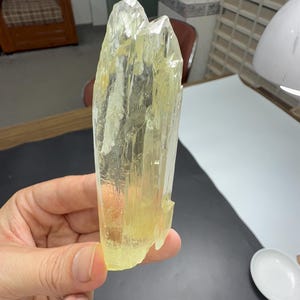 Könnte beinhalten: Ein durchscheinendes, blassgelbes Kristall-Exemplar mit einer doppelt endenden Spitze. Das Mineral weist eine facettierte, längliche Struktur mit natürlichen vertikalen Streifen entlang der Oberfläche auf und zeigt eine klare, glasartige Textur.
