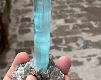 May include: A large, light blue aquamarine crystal with a pointed tip, mounted on a rough, gray rock.