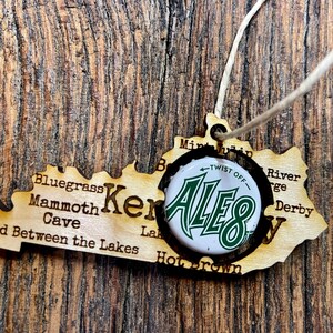 May include: A wooden cutout of the state of Kentucky with a bottle cap featuring the Ale-8-One logo. The cutout includes text such as "Bluegrass", "Mammoth Cave", "Land Between the Lakes", "Mint Julep", "River Gorge", and "Derby".