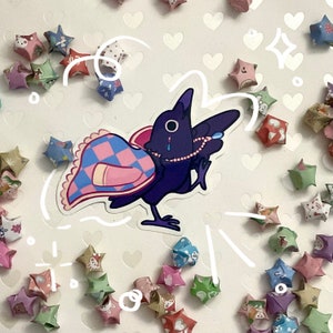 May include: A sticker featuring a purple cartoon crow with a pearl necklace and a patterned blanket. The crow has a tear and is surrounded by origami stars in various colors and patterns, scattered on a white background.