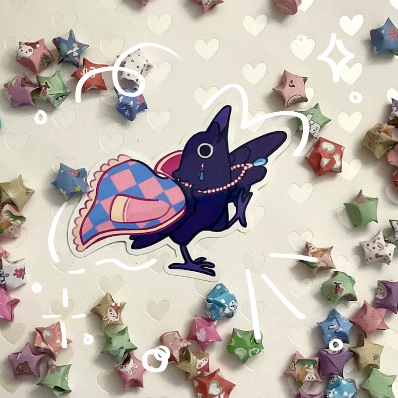 May include: A sticker featuring a purple cartoon crow with a pearl necklace and a patterned blanket. The crow has a tear and is surrounded by origami stars in various colors and patterns, scattered on a white background.