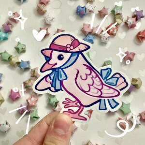 May include: A sticker featuring a cartoon bird in a pink hat and blue scarf. The bird is light pink with purple accents and blue bows. The background is white with heart shapes and origami stars.