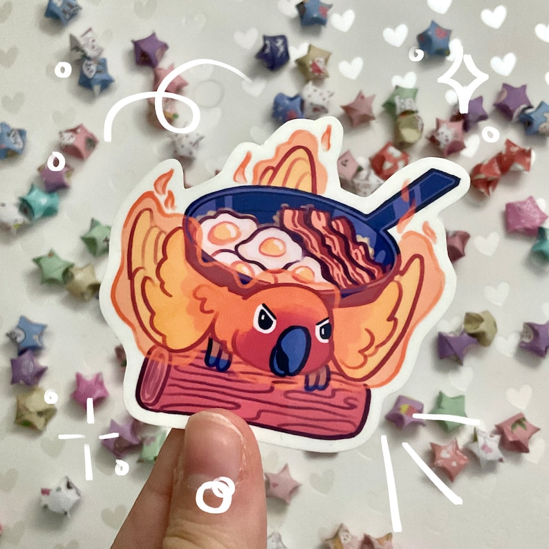 May include: A sticker featuring a cartoon koala with a frying pan on its head, filled with eggs and bacon. The koala is surrounded by flames and perched on a log. The sticker has a white border and is set against a background of origami stars.
