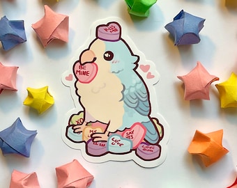 May include: A sticker featuring a cartoon parrot with blue and cream-colored feathers, wearing a candy heart hat that says "Kiss Me." The parrot is surrounded by candy hearts with phrases like "Be Mine" and "Say Yes." Origami stars in various colors surround the sticker.